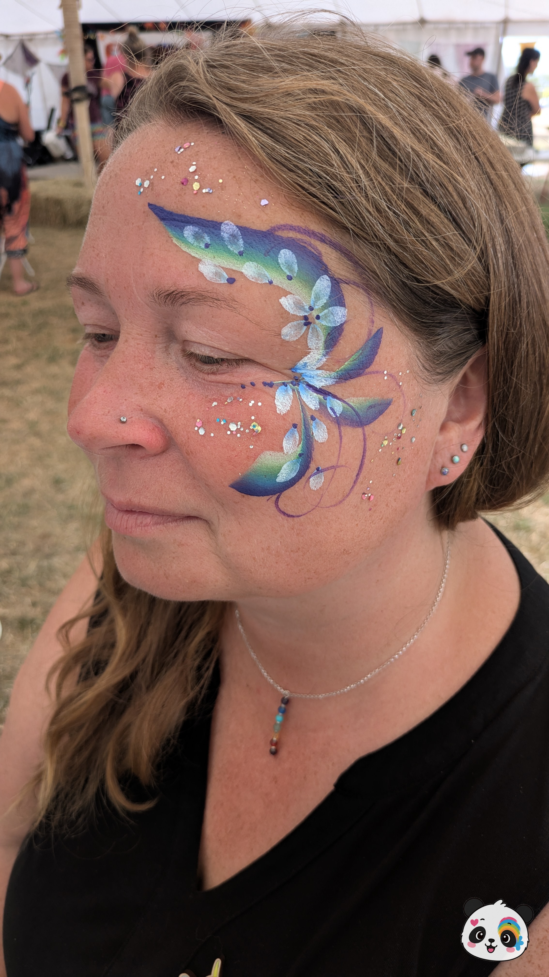 Blue, green, and purple floral face paint with white highlights and glitter accents extending from the forehead to the cheek.