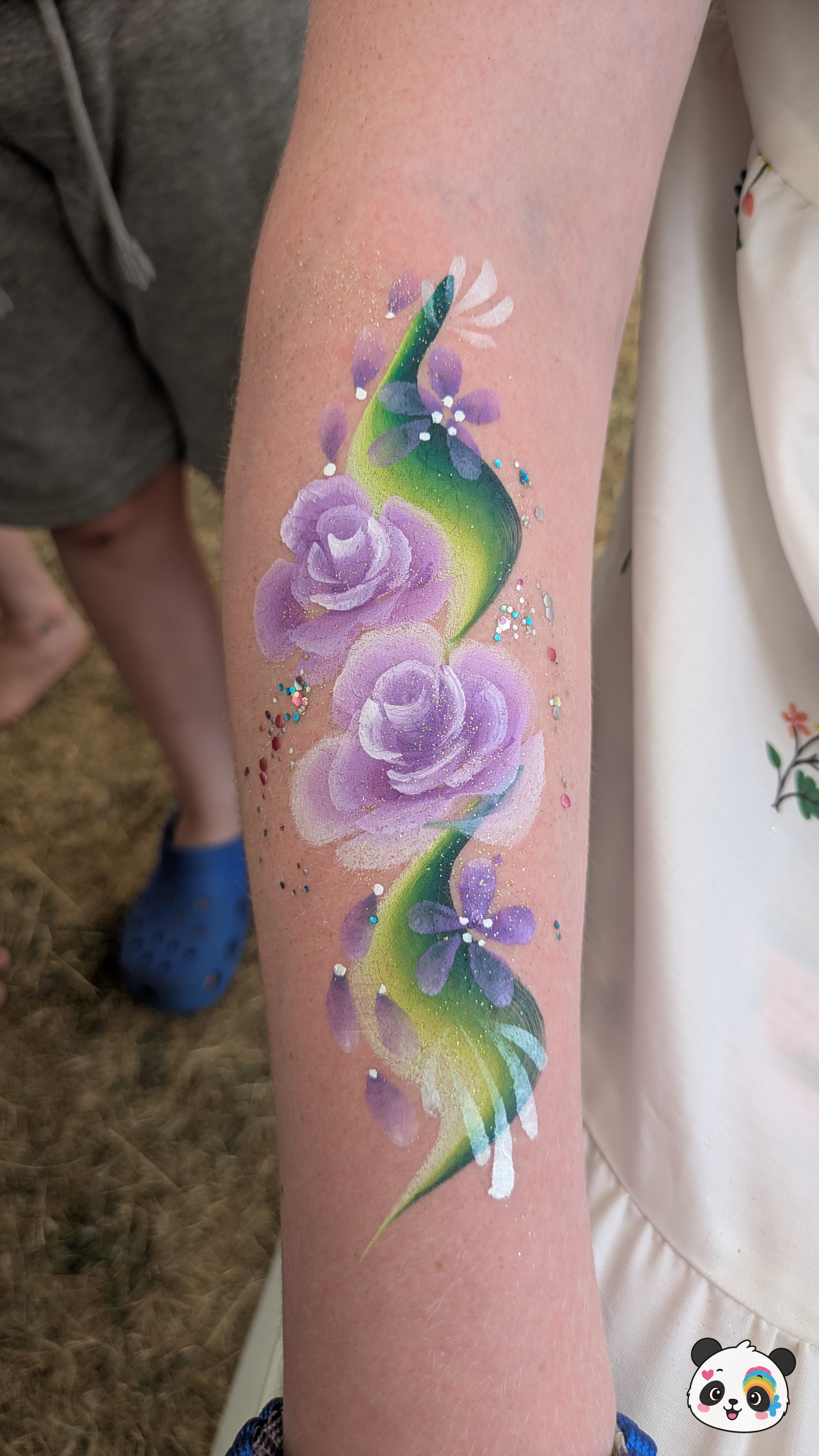 Purple roses and small flowers on a green vine painted on an arm with glitter accents.