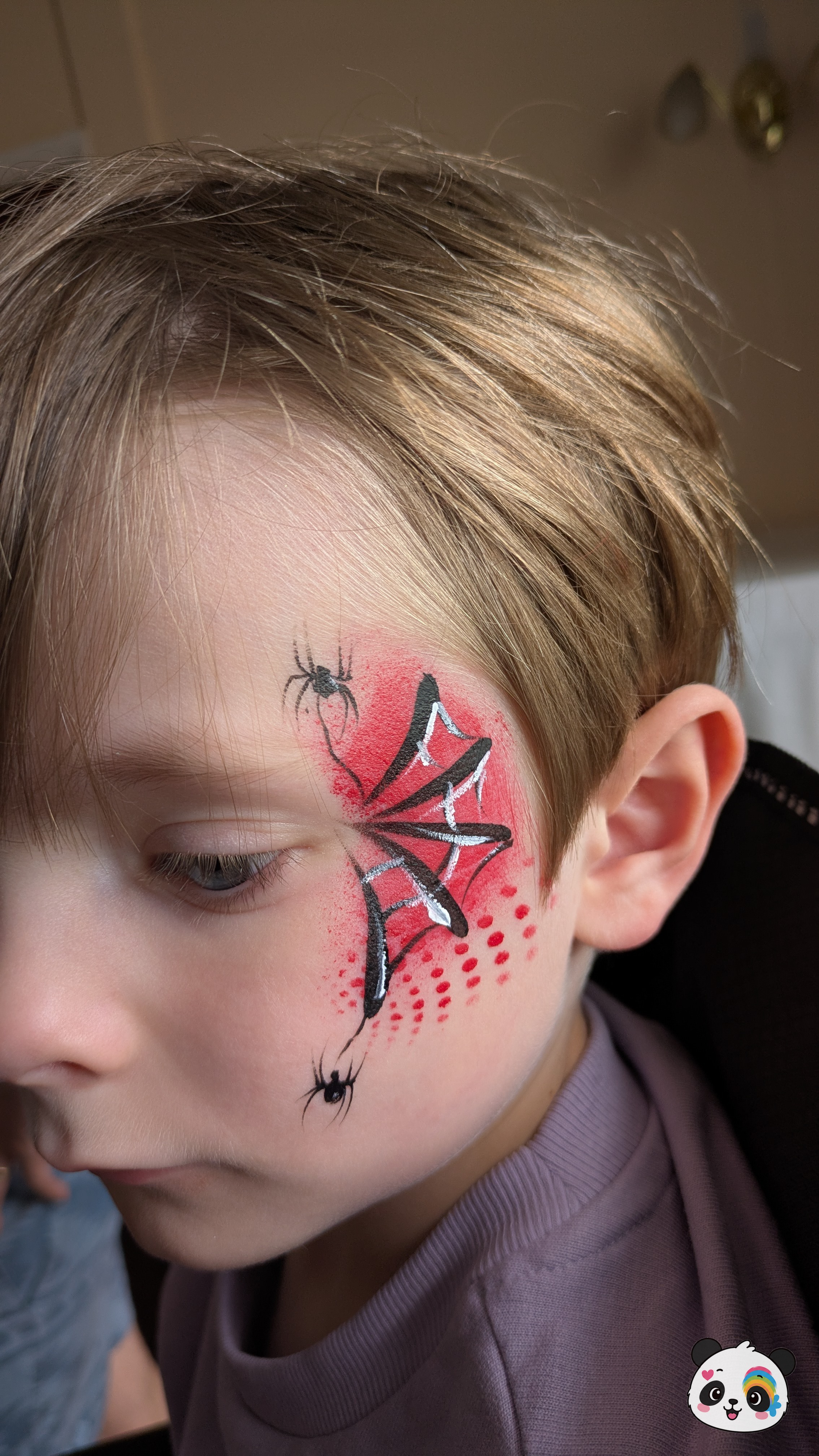Red and black spider web face paint with silver highlights and two small spiders on the temple and cheek.