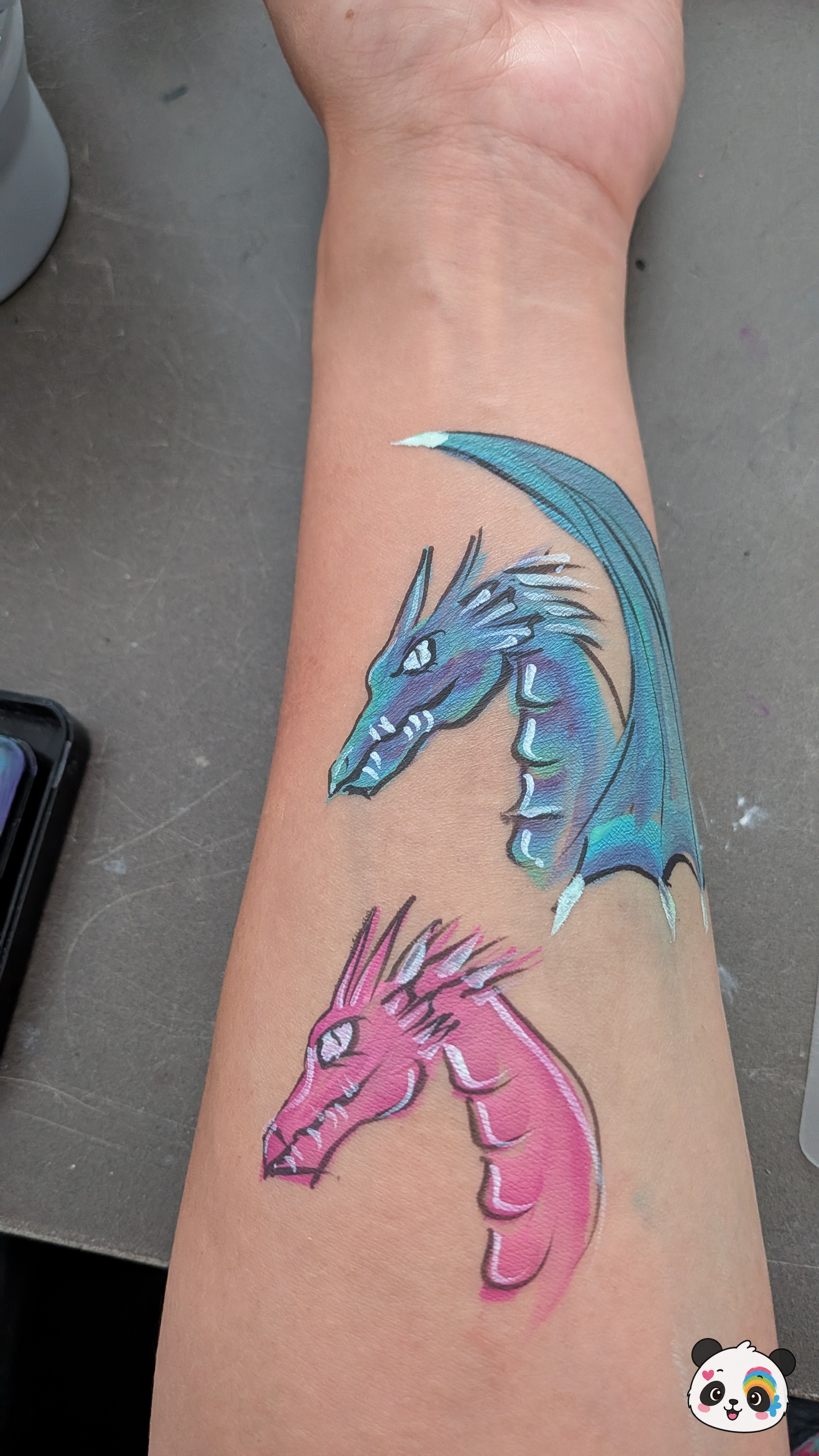 A blue and a pink dragon head painted on an arm with white highlights and black outlines.