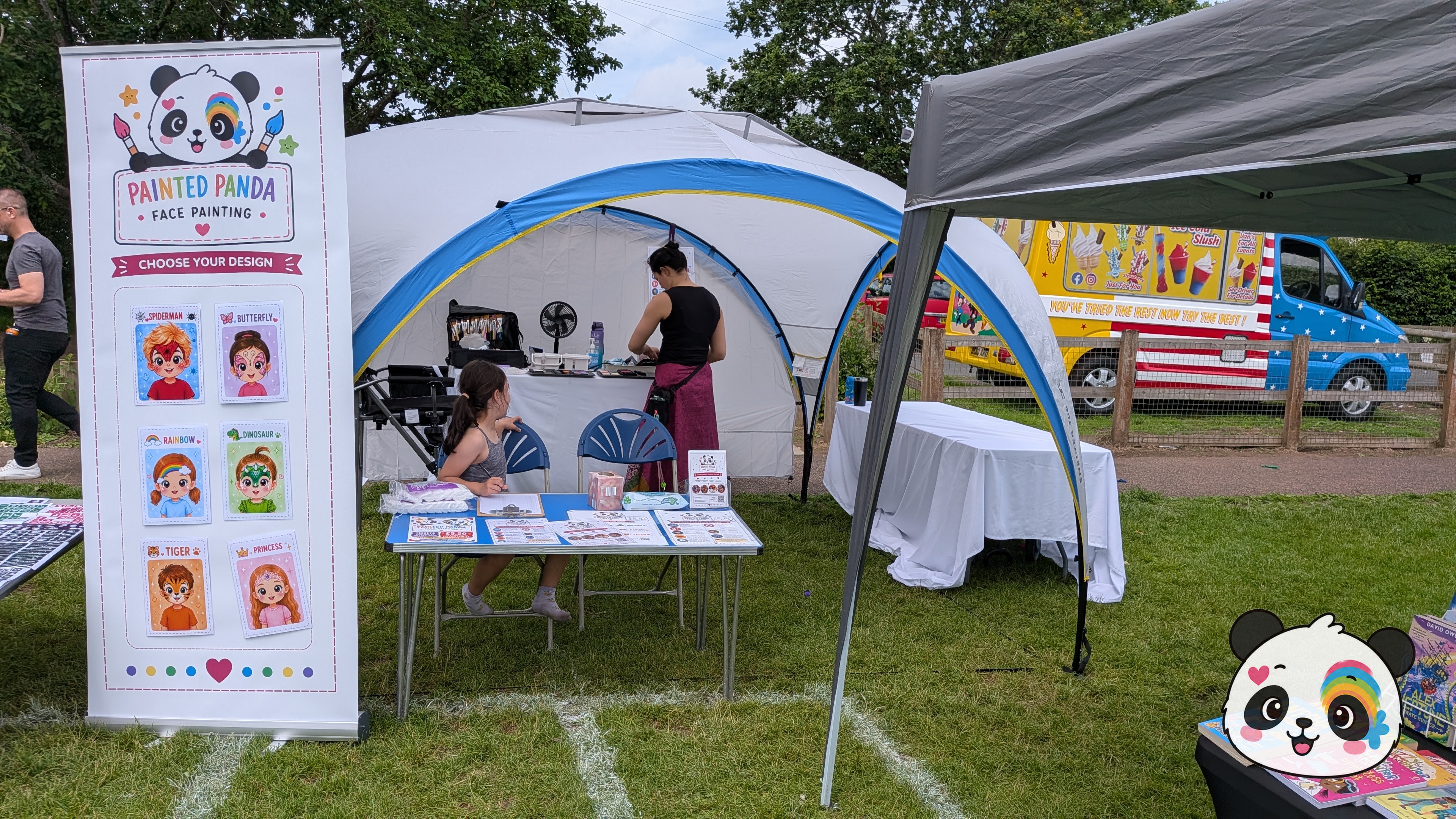 A professional face painting setup featuring a branded banner with design examples, a white canopy tent, and a table with supplies on a grassy field.