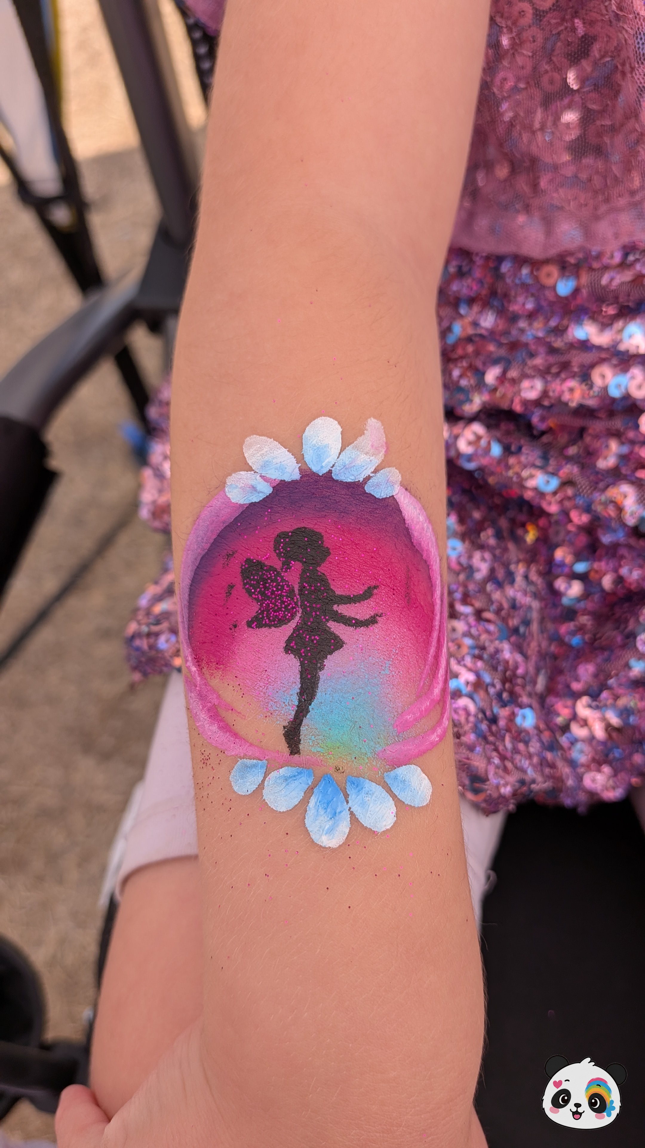 A black fairy silhouette painted inside a pink, purple, and blue circular background on an arm, framed by light blue flower petals.