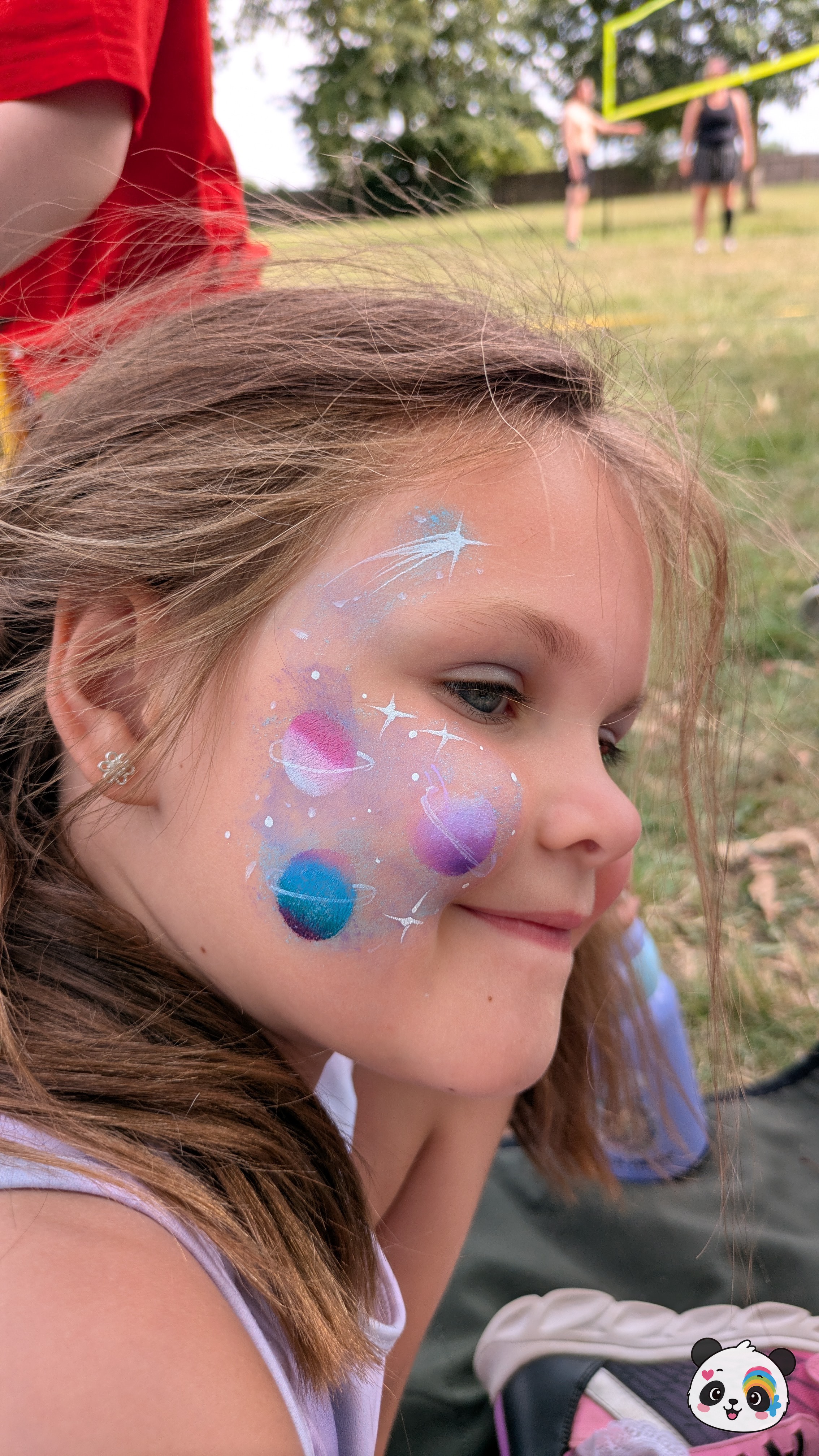 Outer space themed face paint featuring planets, stars, and a shooting star in shades of blue, purple, and pink on the cheek and temple.