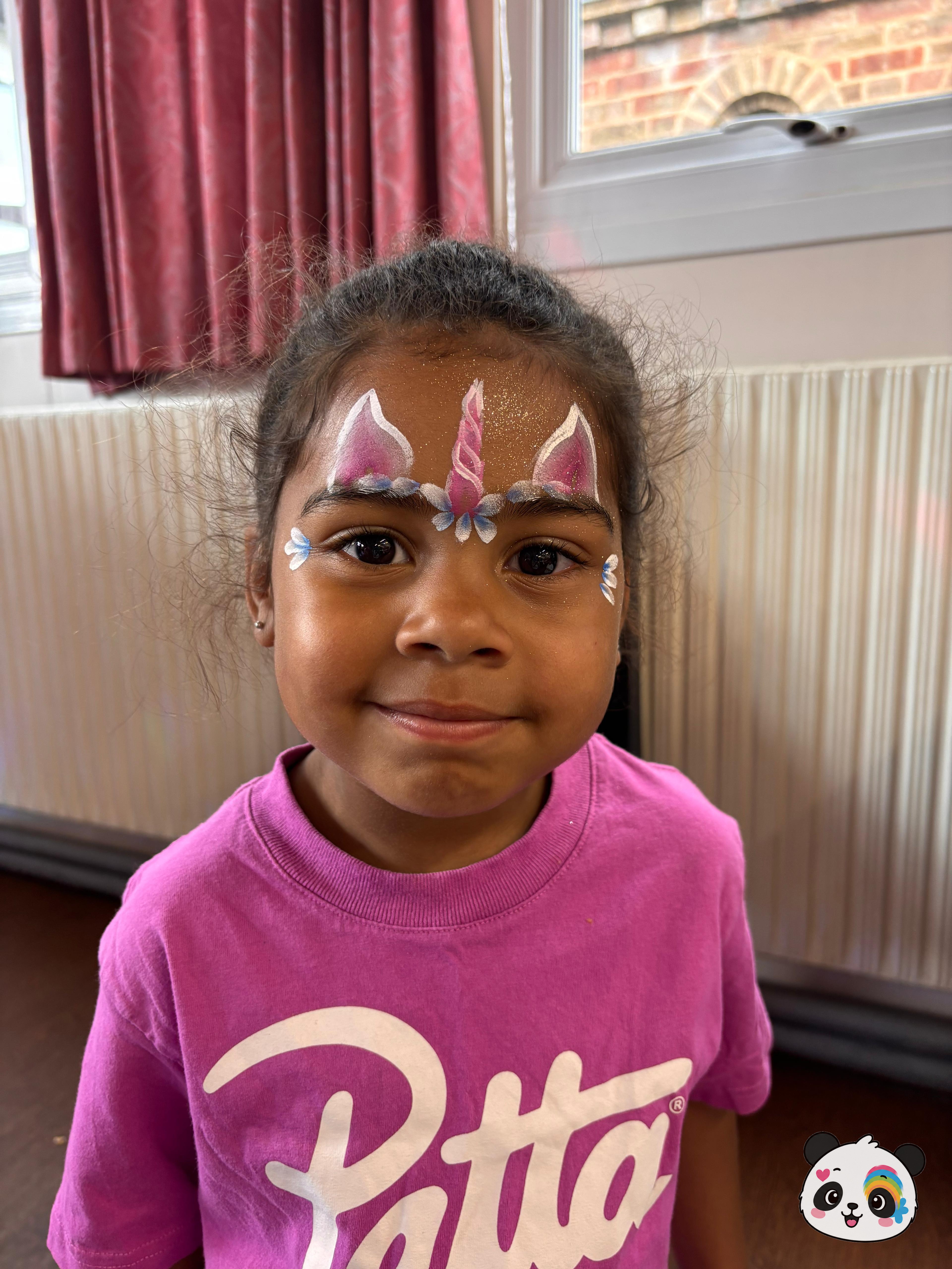 Pink and white unicorn horn and ears painted on the forehead with small blue flower accents near the eyes.