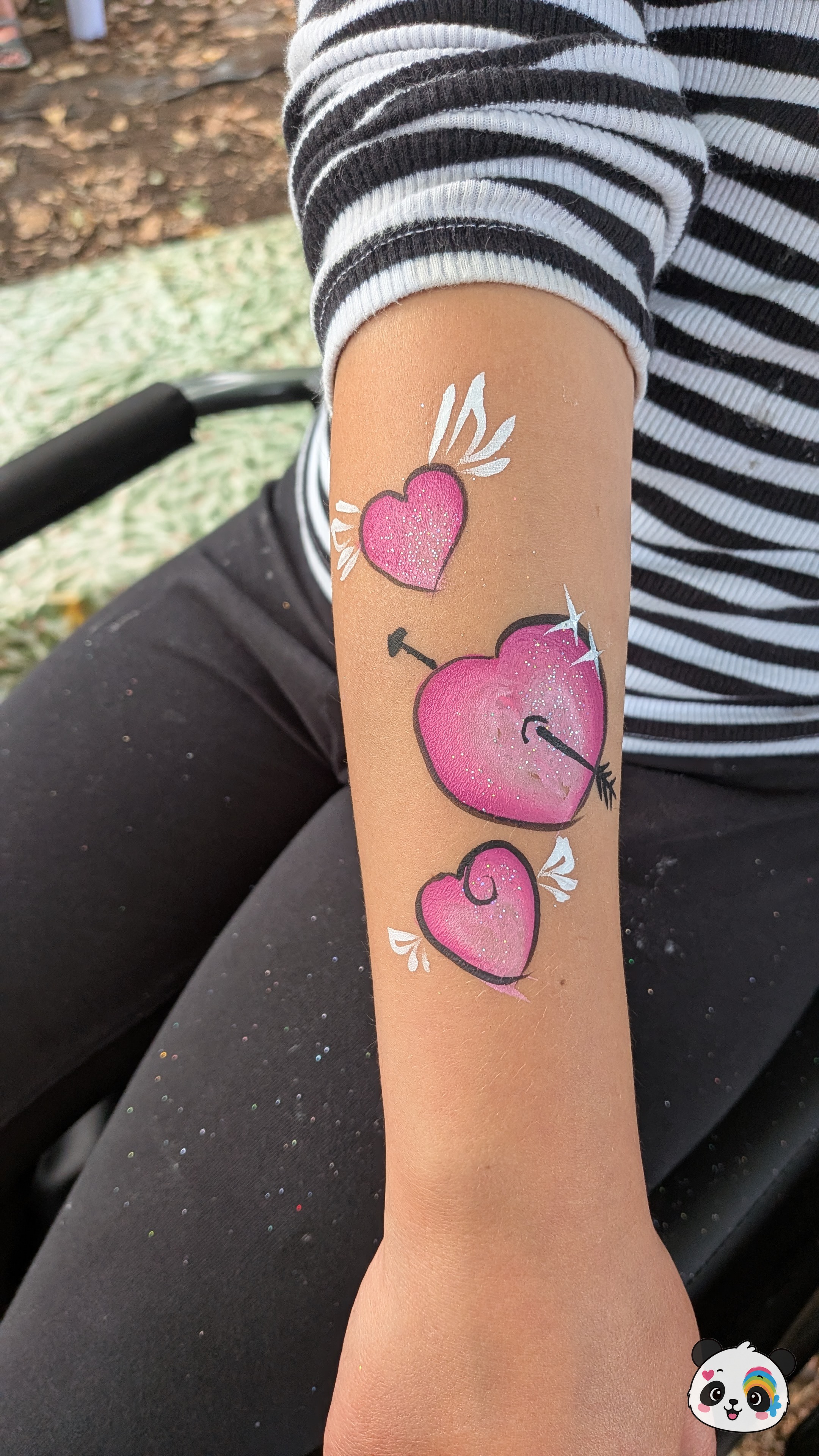 Three pink glittery hearts with white wings and a black arrow design painted on a forearm.