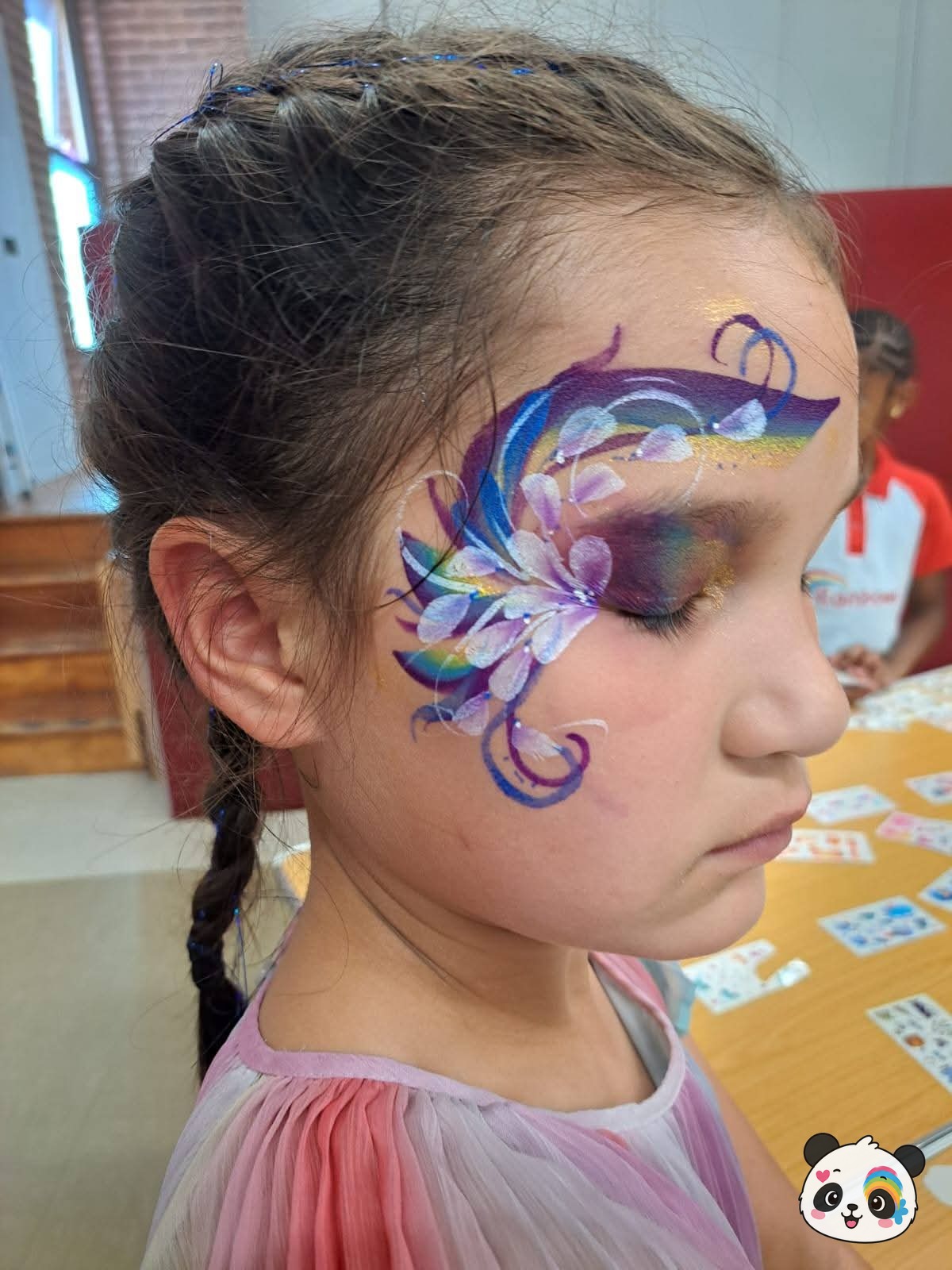 Rainbow and purple floral face paint design with white highlights extending from the temple across the cheek.