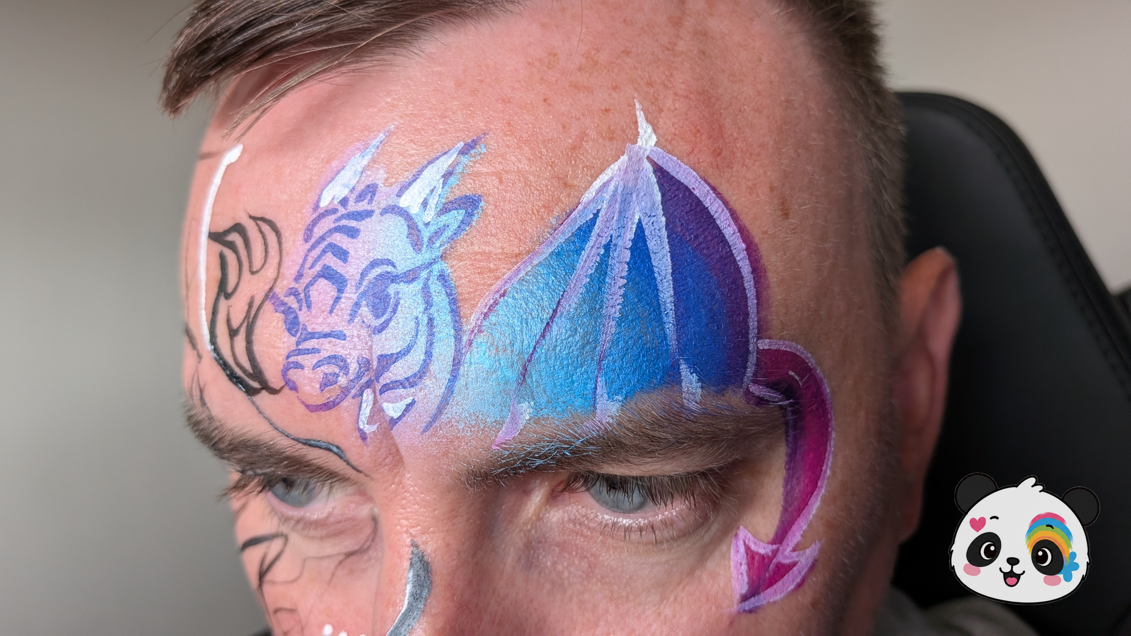 Blue and purple dragon face paint featuring a dragon head and wings on the forehead and temple area.