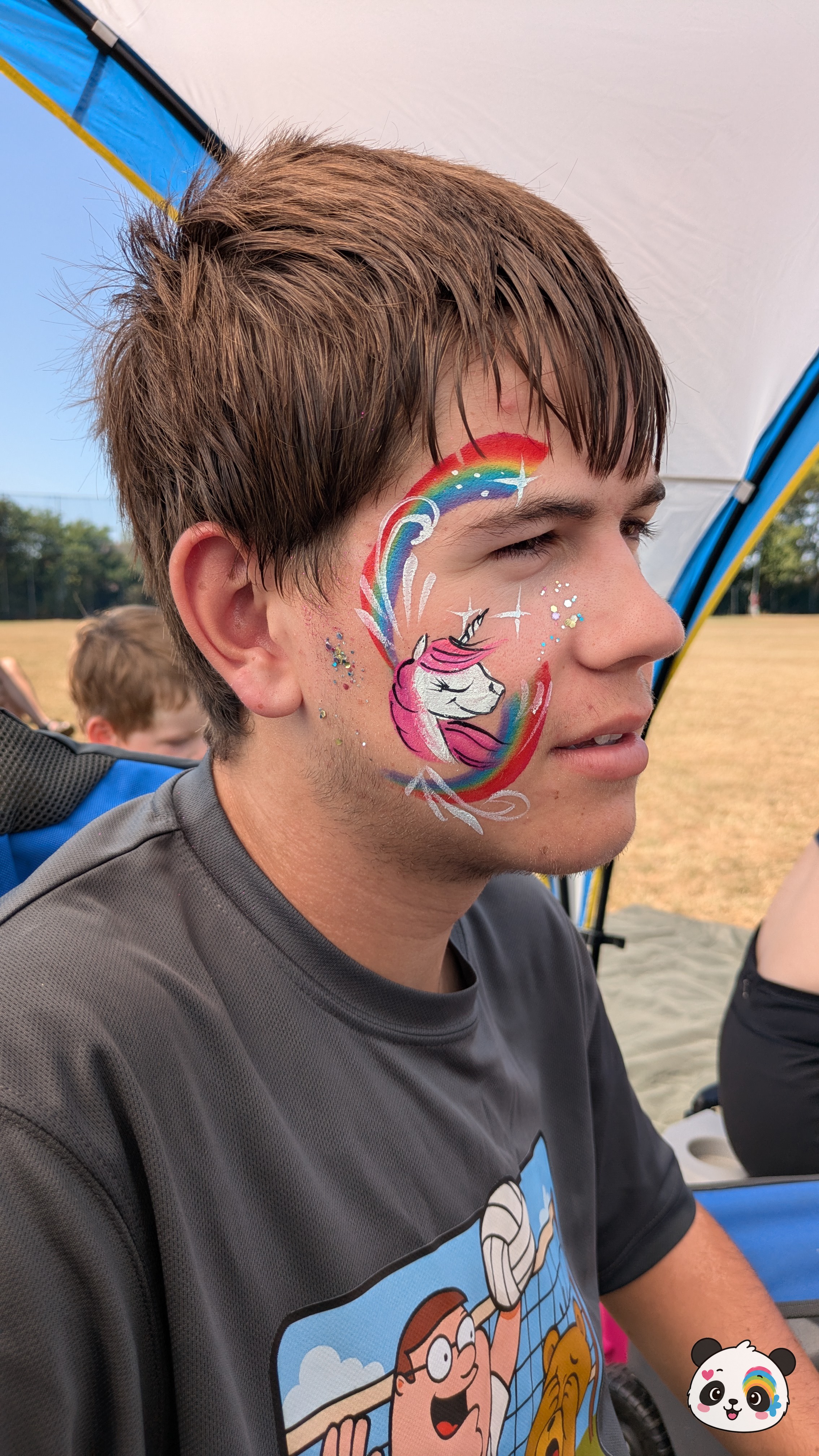 A pink and white unicorn face paint design accented with a vibrant rainbow arch, white star details, and glitter on the cheek and temple.