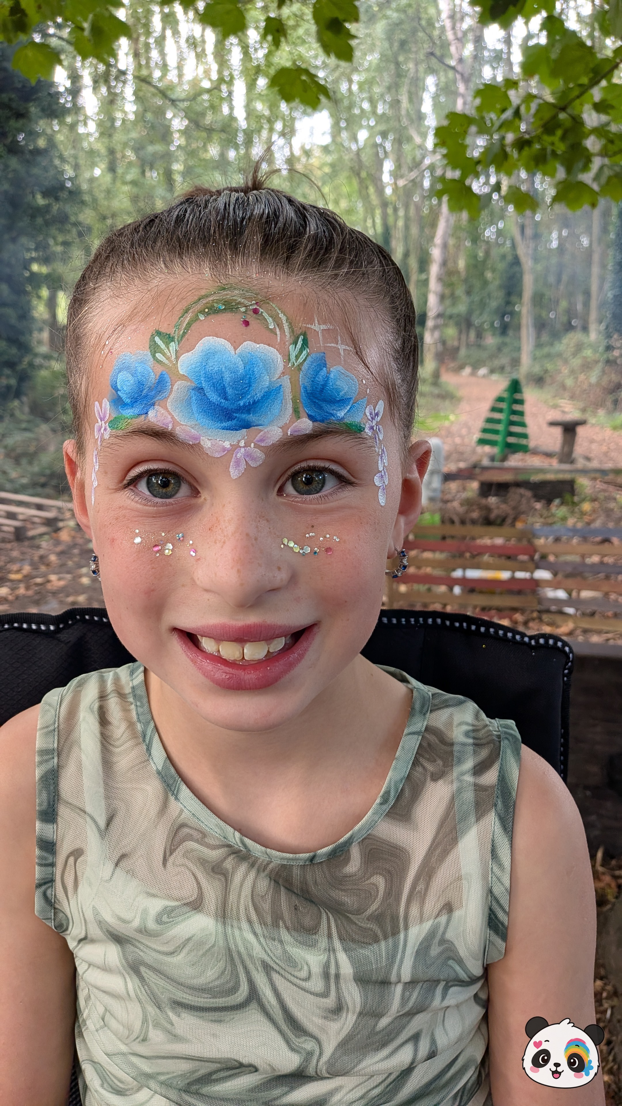 Blue rose face paint design with green leaves and white floral accents across the forehead, finished with glitter on the cheeks.