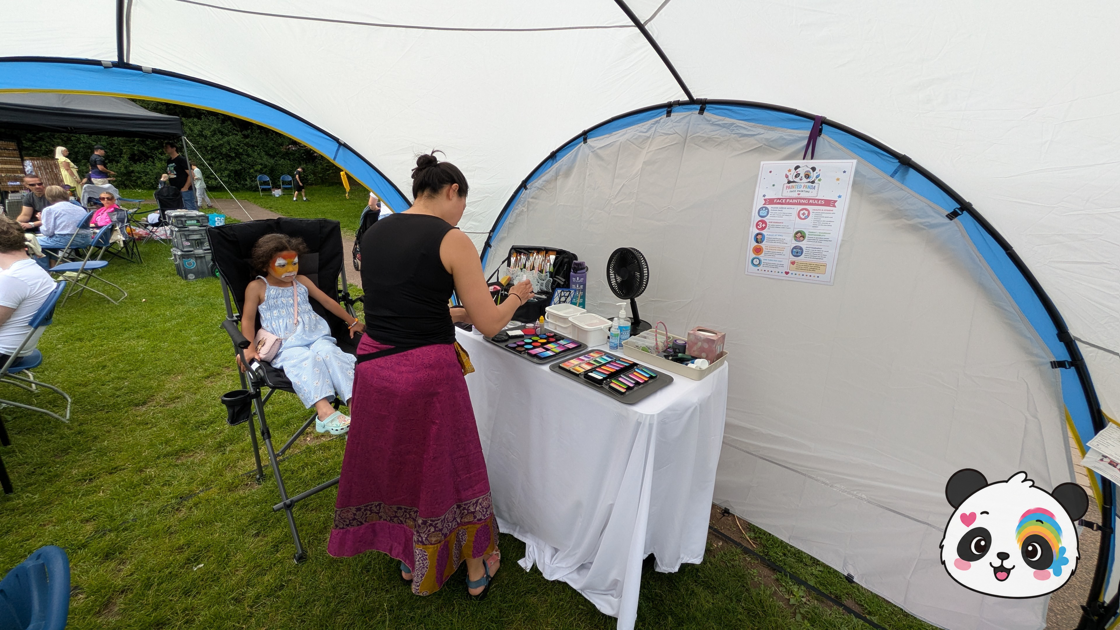 Orange, yellow, and black tiger face paint design on a child's face while an artist works at a pop-up station.