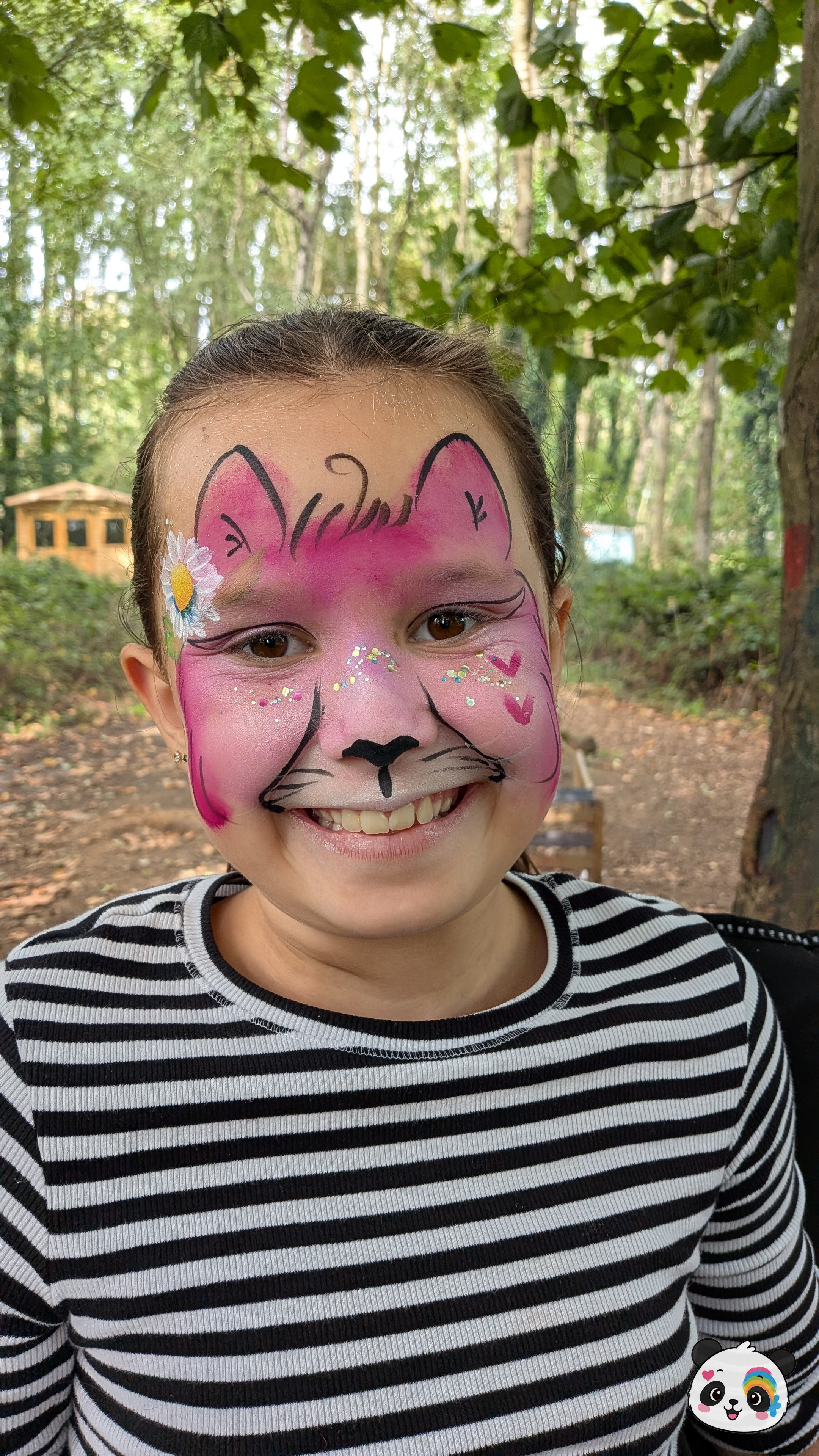 Pink and white cat face paint with black whiskers, glitter accents, and a small daisy flower on the temple.