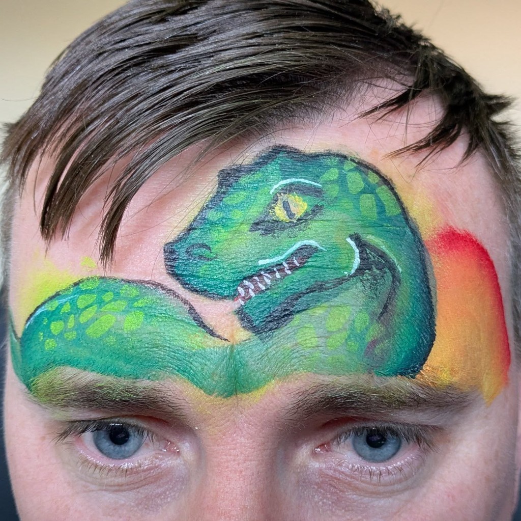 Green dinosaur face paint across a child's forehead with sunset background