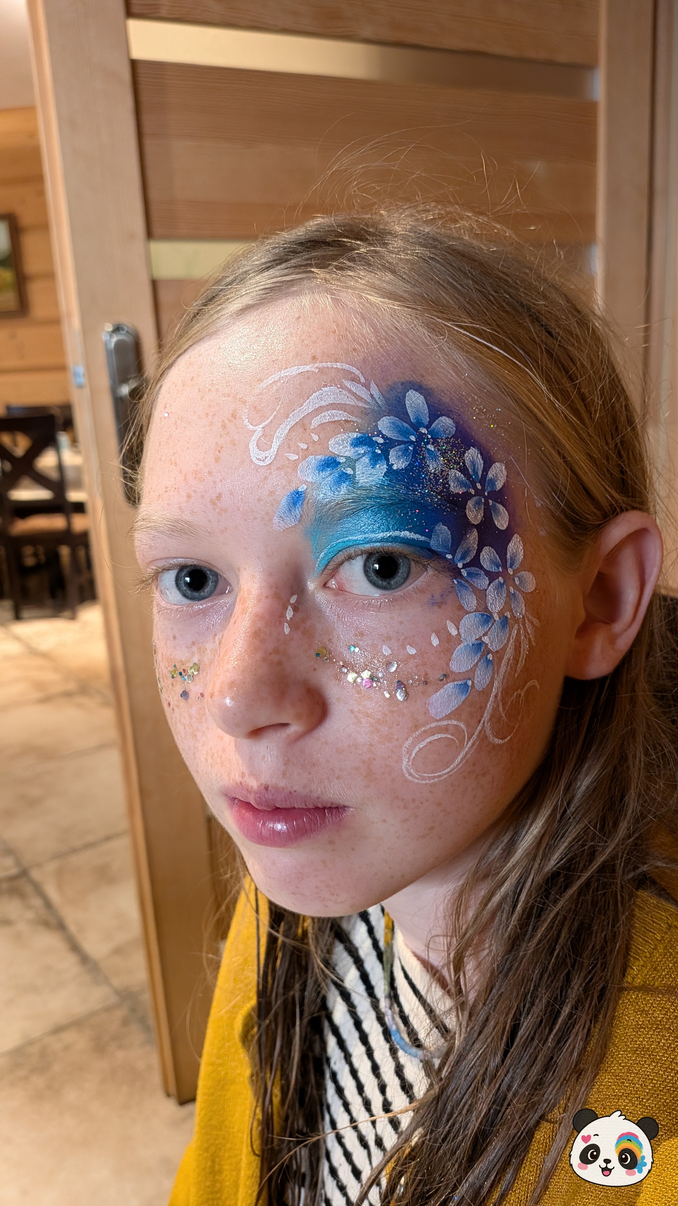 Blue and purple floral face paint with white swirls and glitter accents on the forehead and cheek.
