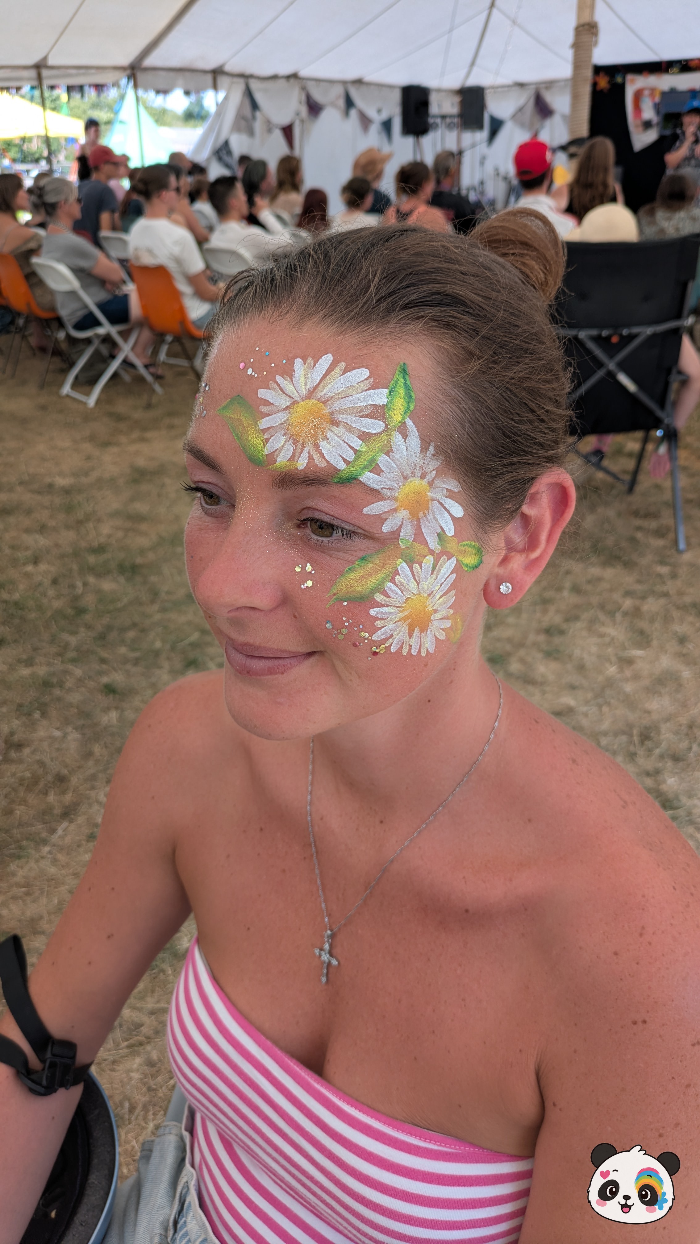 White and yellow daisy flower face paint with green leaves on the forehead and cheek, accented with glitter.